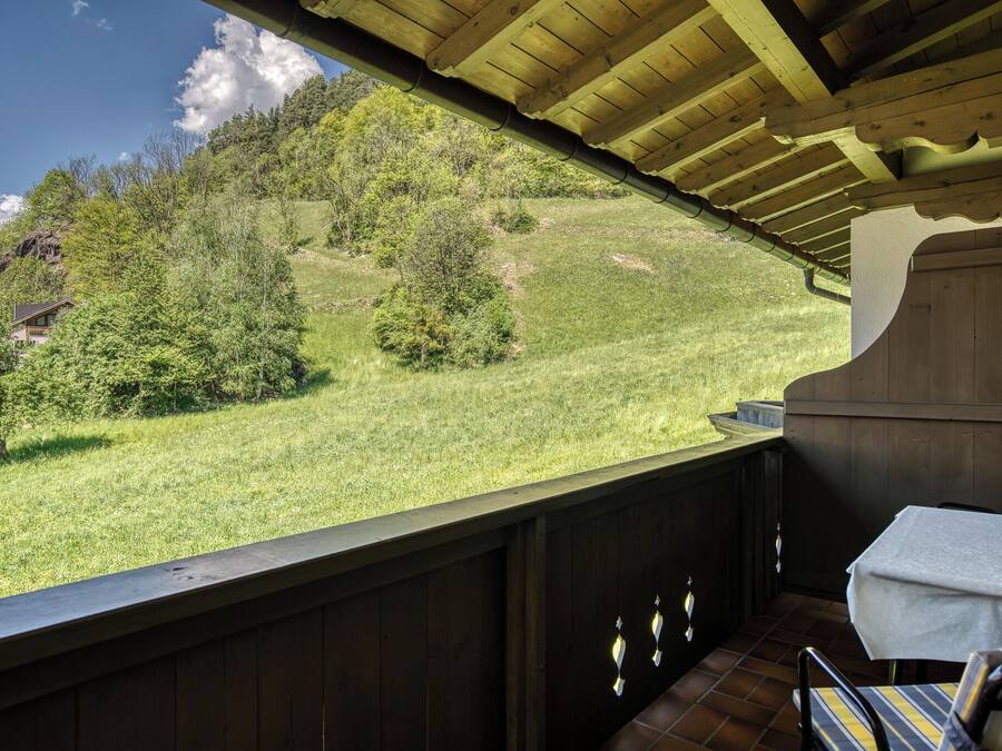 Una terraza de madera con una mesa y sillas, con vistas a una ladera cubierta de césped, árboles y una casa a lo lejos.
