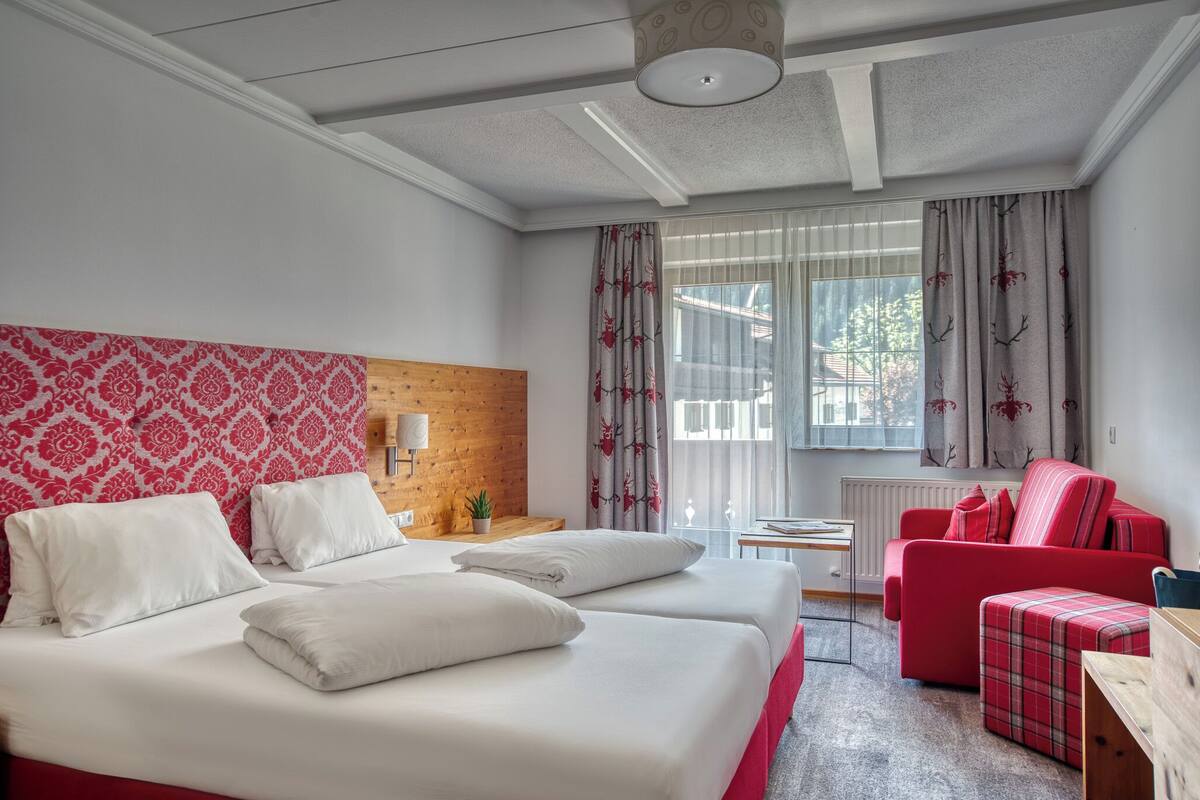 A hotel room with two beds, a red sofa, a plaid ottoman, a wooden headboard, and a window with curtains.