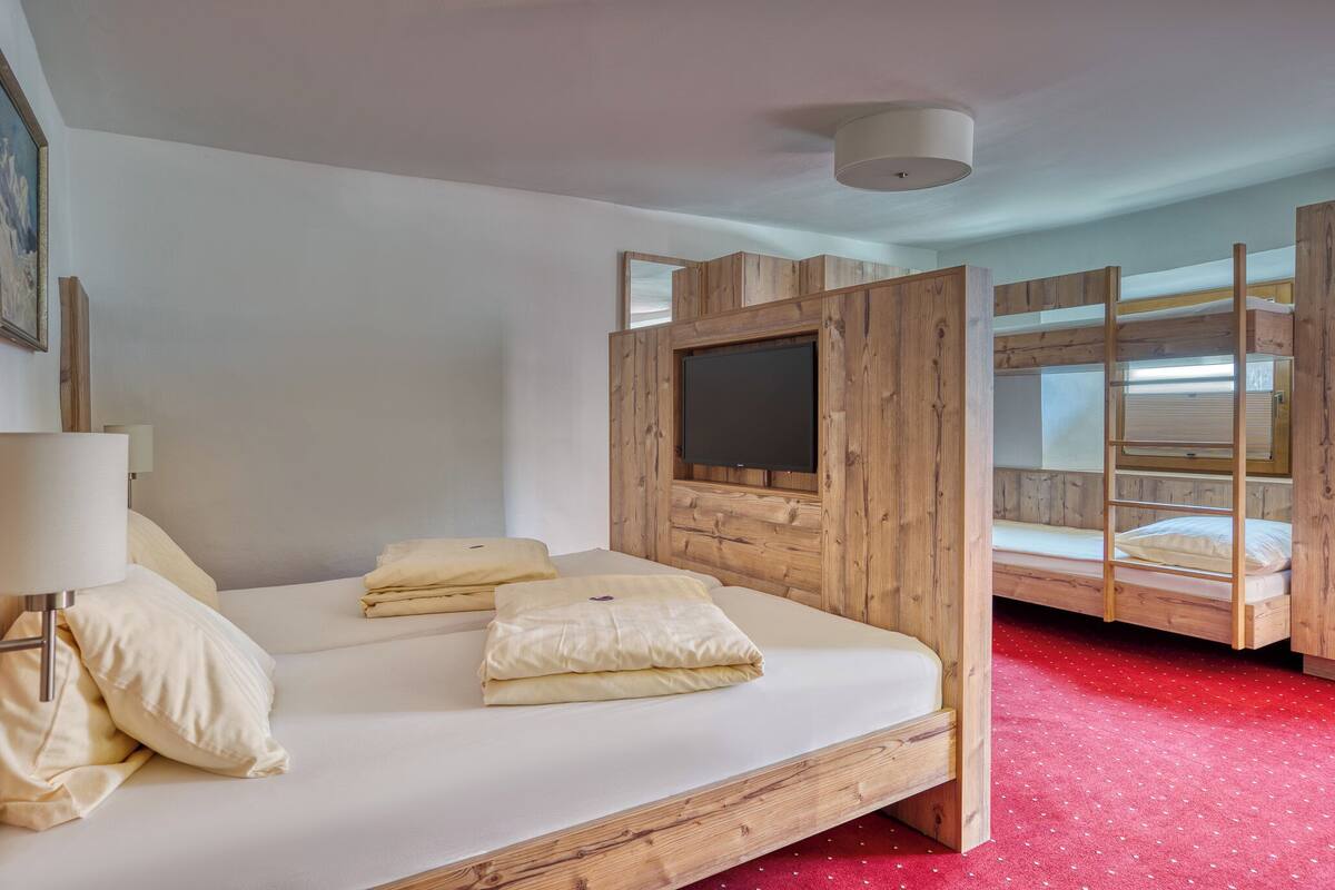 A room with a single bed, a TV mounted on a wooden panel, and a bunk bed in the background.