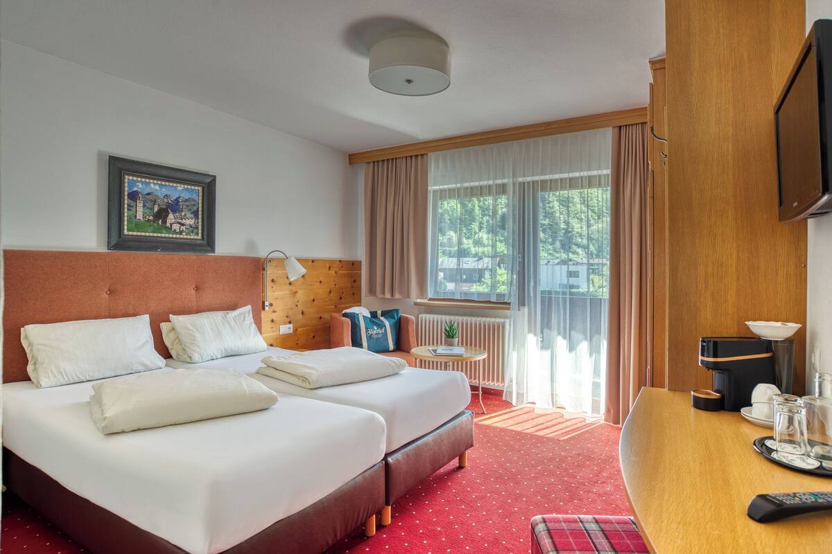 A hotel room with two beds, a small table with a lamp, a TV, and a window with curtains.