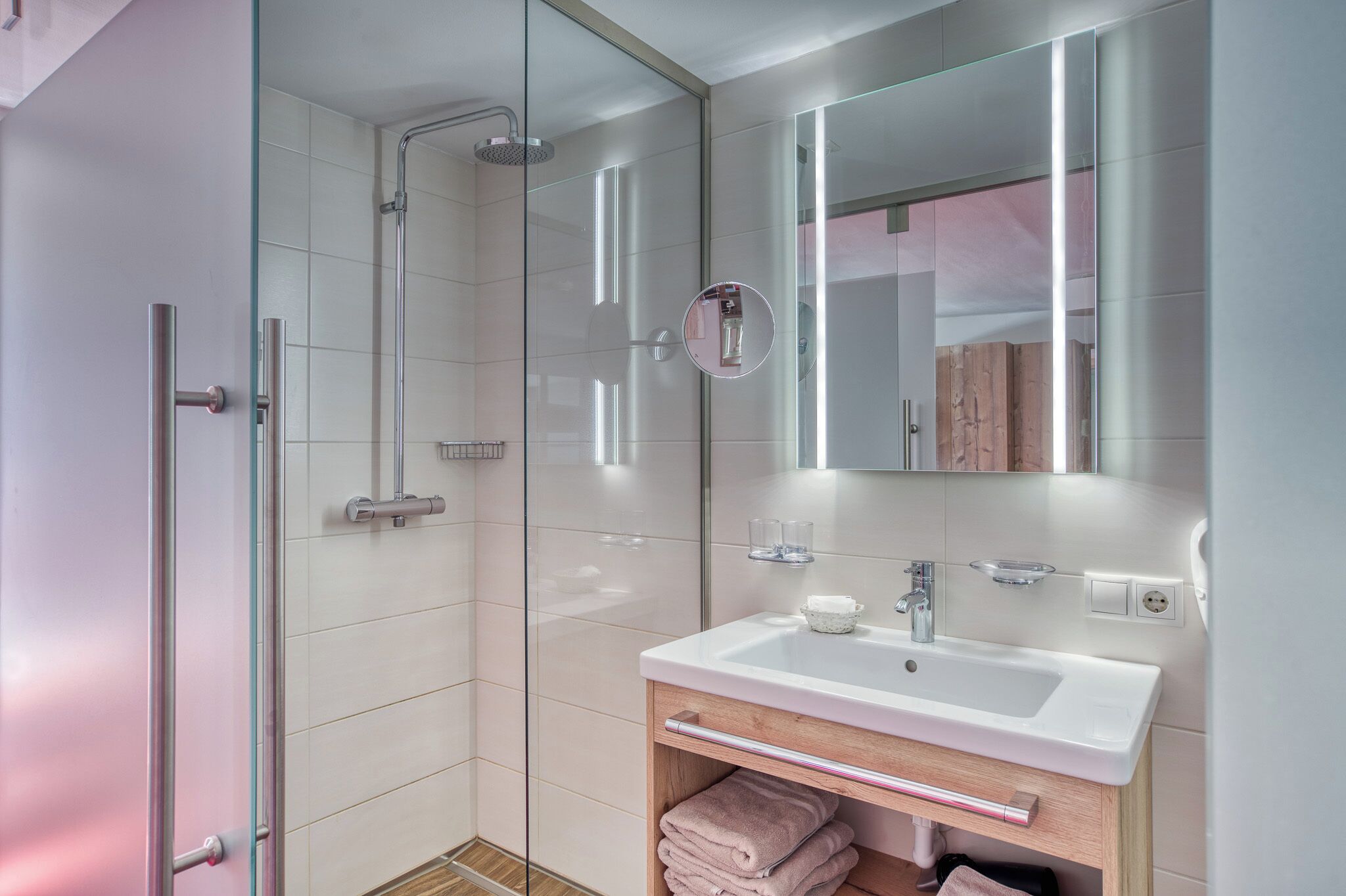 A modern bathroom with a glass-enclosed shower, a white sink with a wooden vanity, and a mirror with integrated lighting.