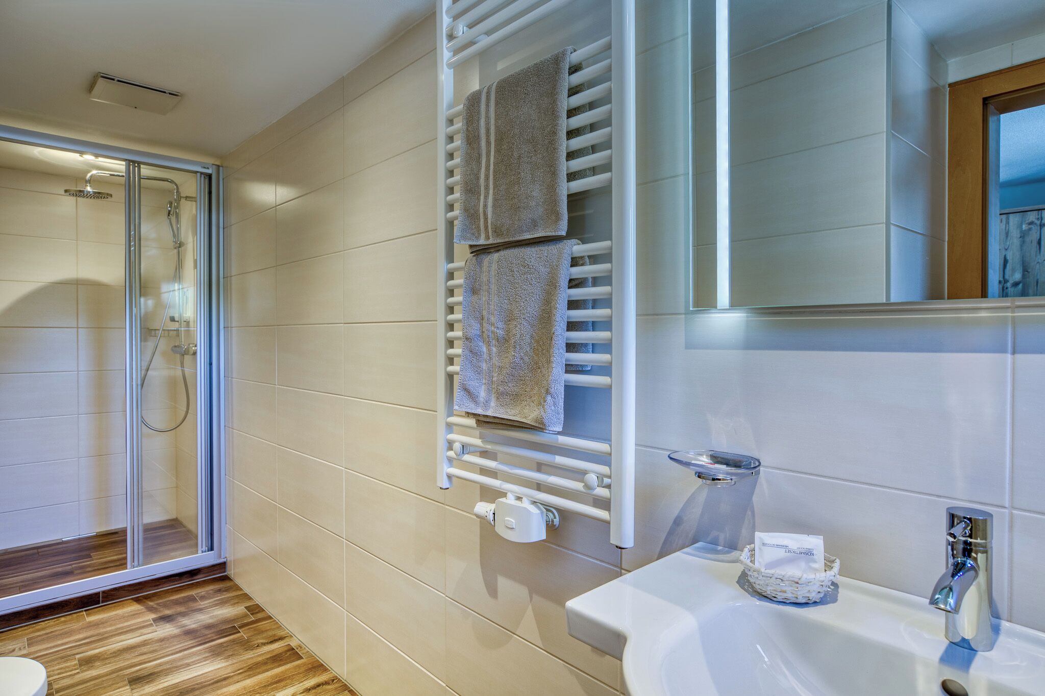 Un baño moderno con cabina de ducha de vidrio, un perchero con dos toallas, un lavabo blanco y un espejo.