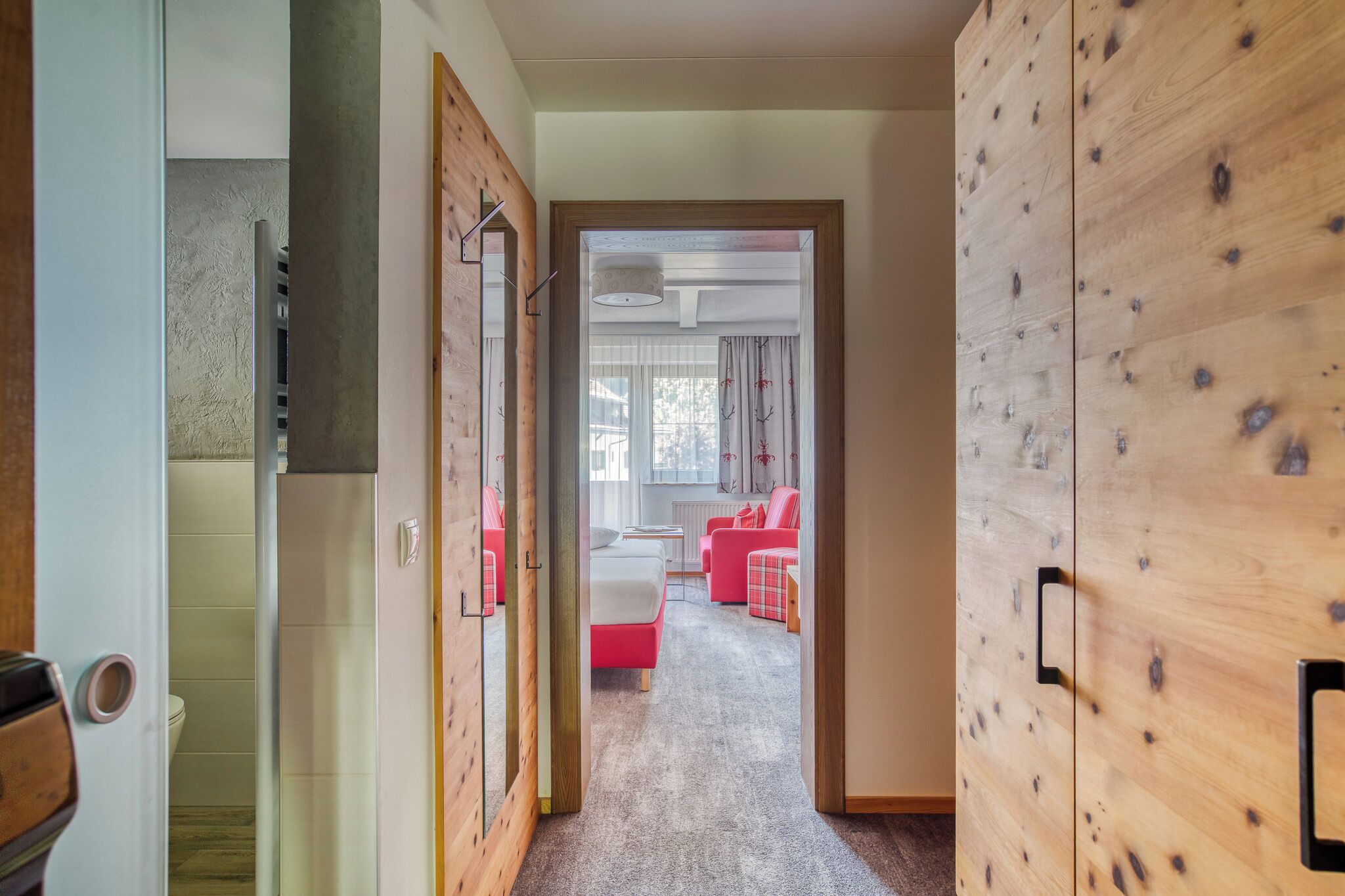 Ein Flur, der zu einem Schlafzimmer führt. Im Zimmer befindet sich ein Bett, ein Schreibtisch und ein Stuhl. Rechts ist ein Holzschrank und im Hintergrund ist ein Badezimmer mit Toilette zu sehen.