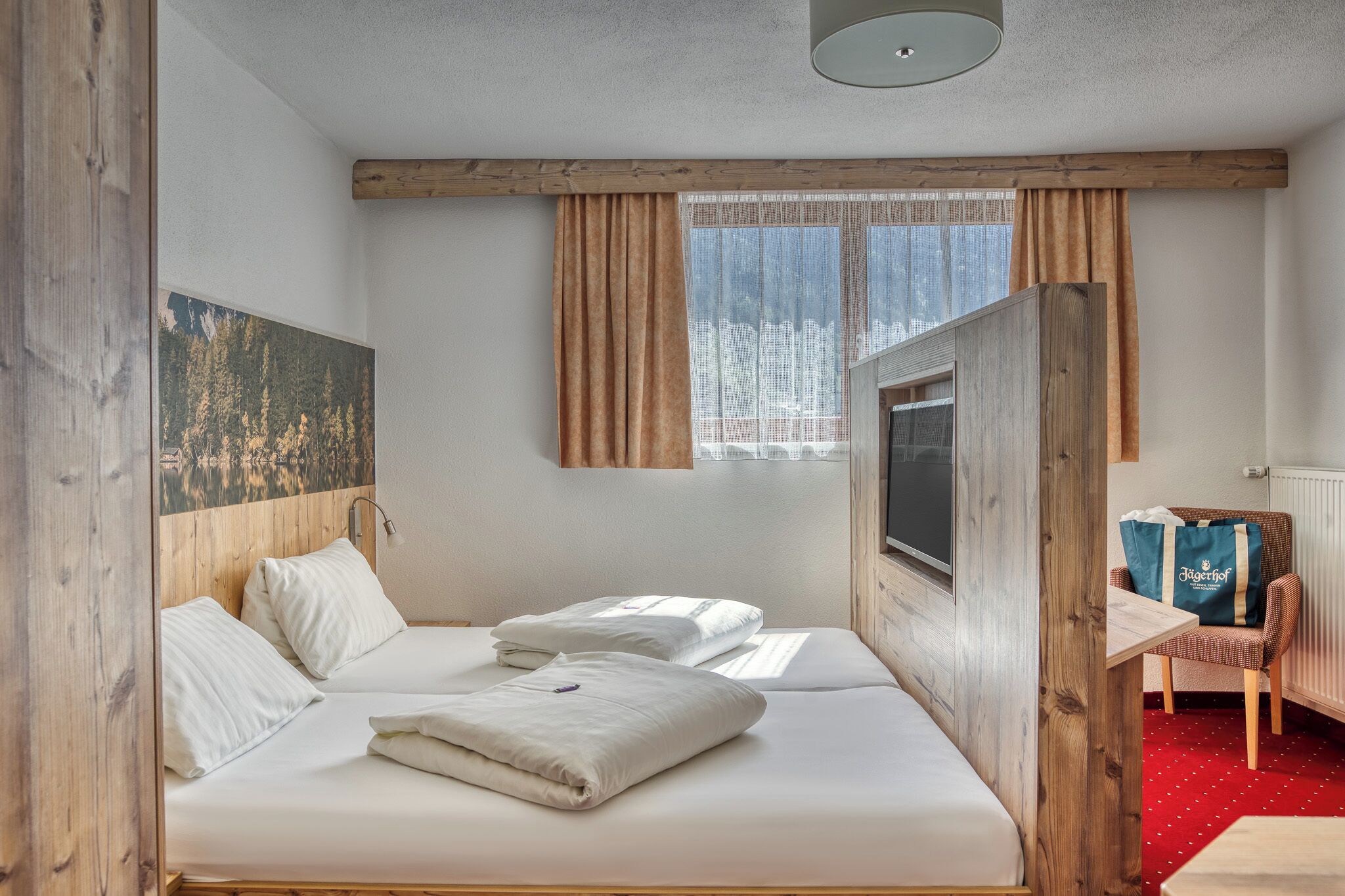 Habitación de hotel con cabecera de madera, dos camas, un televisor, una ventana con cortinas, una alfombra roja y una pequeña mesa con una silla.
