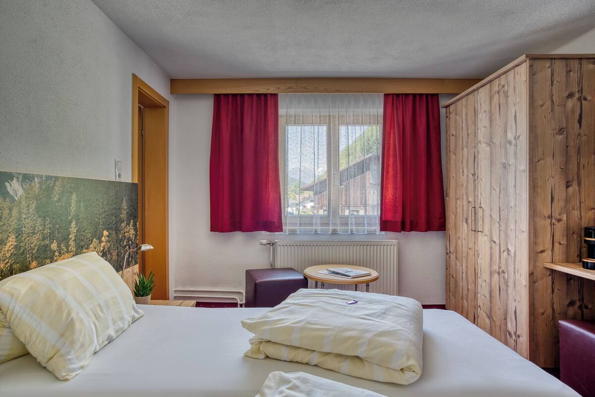 A hotel room with a bed, a small table, a chair, a wooden wardrobe, and a window with red curtains.