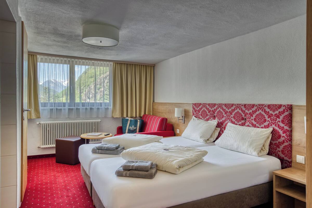 A hotel room with a large bed, two bedside tables, a red and white patterned headboard, a window with curtains, and a radiator.