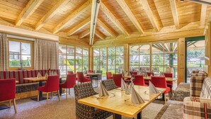 Ein Restaurant mit Holzbalken, rot-gemusterten Stühlen und großen Fenstern mit Blick ins Freie.