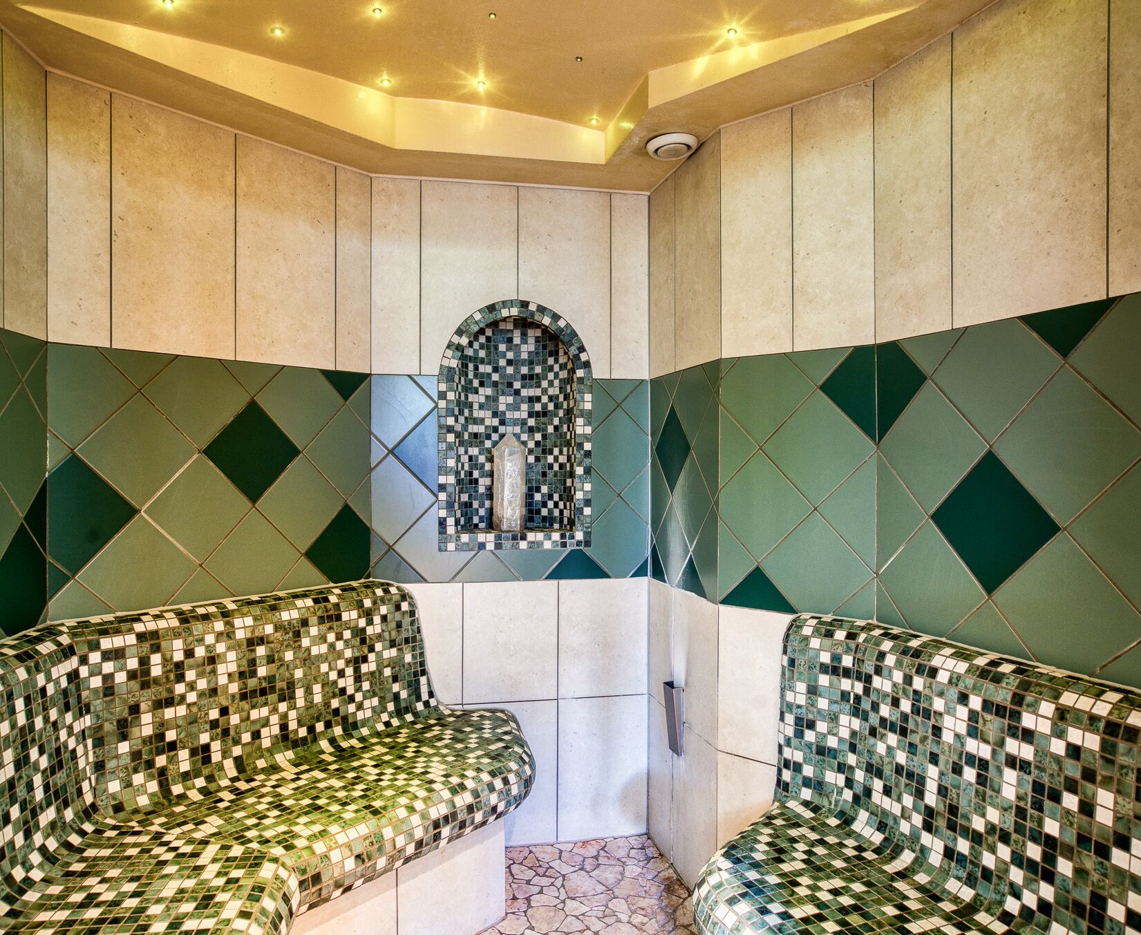 A spa room with two mosaic-patterned benches, a tiled wall with a mosaic arch, and a stone floor.