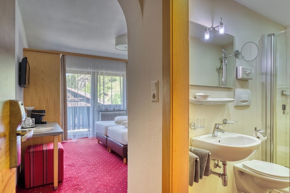 A hotel room with a bed, a desk, a suitcase, a red carpet, a bathroom with a sink, mirror, and shower. - Hotel Jägerhof (Oetz)