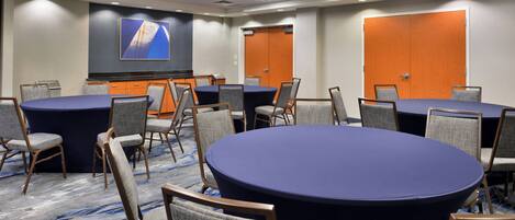 Une salle de conférence équipée de tables rondes, de chaises, d’un écran de projection et d’un sol recouvert de moquette.