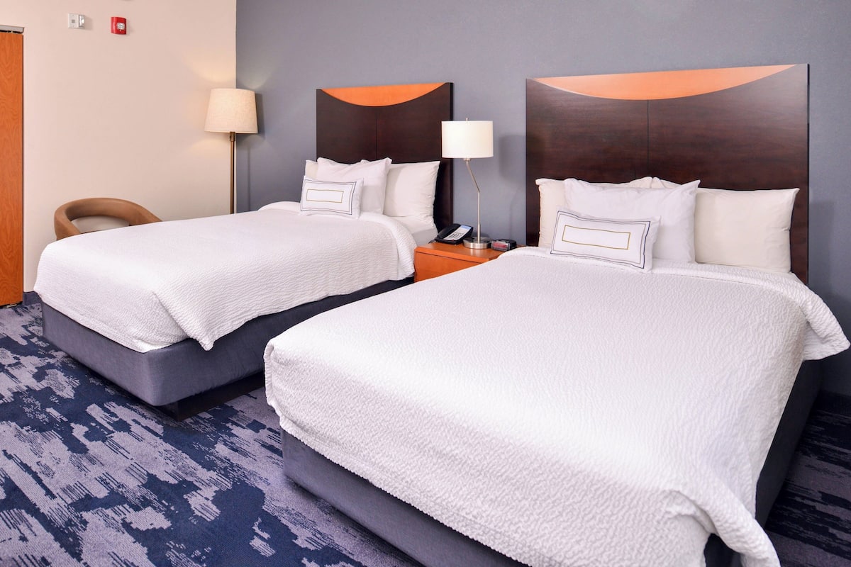 A hotel room with two beds, a nightstand with a lamp, and a chair.