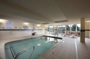 An indoor swimming pool with a lifeguard on duty sign, lounge chairs, and a view of the outdoors.