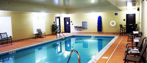 An indoor swimming pool with a clear water surface, a metal ladder, and a tiled edge. There are chairs and a table with papers on the right side.