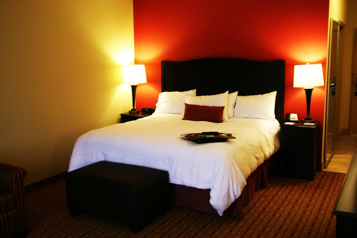 Ein Hotelzimmer mit einem großen Bett, zwei Nachttischen mit Lampen, einer kleinen Bank und Teppichboden.
