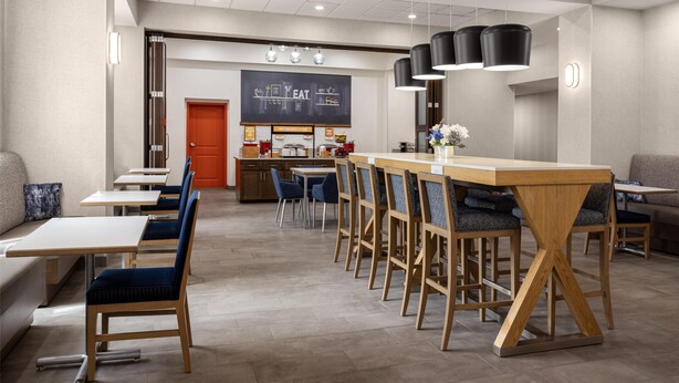 Un comedor moderno con mesas y sillas de madera, una cocina al fondo e iluminación colgante.