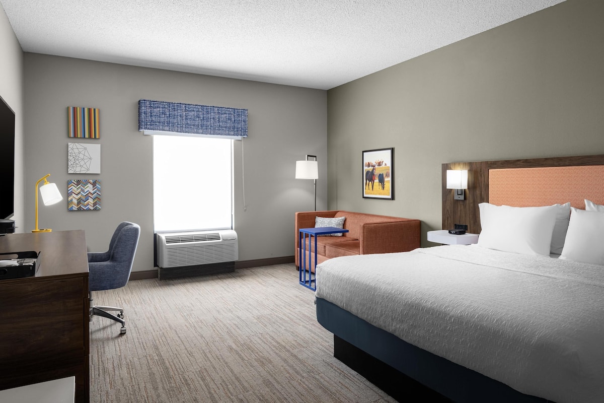 Ein Hotelzimmer mit einem großen Bett, einem Schreibtisch mit Stuhl, einem kleinen Tisch und einem Fenster mit blauer Vorhangbesatz.