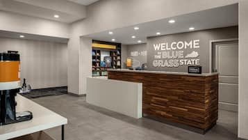 A reception area with a wooden reception desk, a coffee machine, and a wall sign welcoming guests to Blue Grass State.