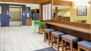 Un hall d’hôtel avec un comptoir d’accueil, des tabourets en bois, une plante en pot et un ascenseur.
