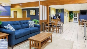 Un hall d’hôtel avec un canapé bleu, des tables et des chaises en bois. On y trouve un comptoir d’accueil, une machine à café et un téléviseur fixé au mur.