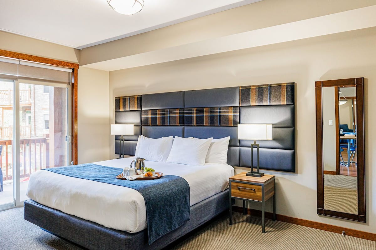 A hotel room with a large bed, a nightstand with a lamp, a mirror, and a view of the outside balcony.