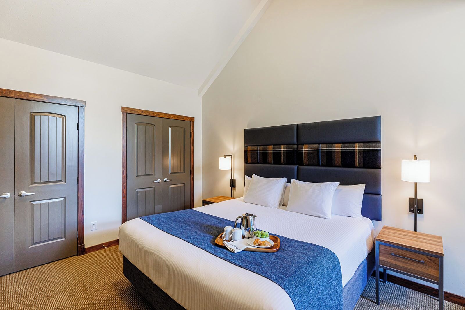 A hotel room with a large bed, a wooden nightstand, two bedside lamps, and a tray with breakfast items on the bed.