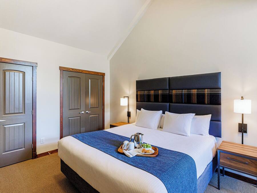 A hotel room with a large bed, a wooden nightstand, two bedside lamps, and a tray with breakfast items on the bed.