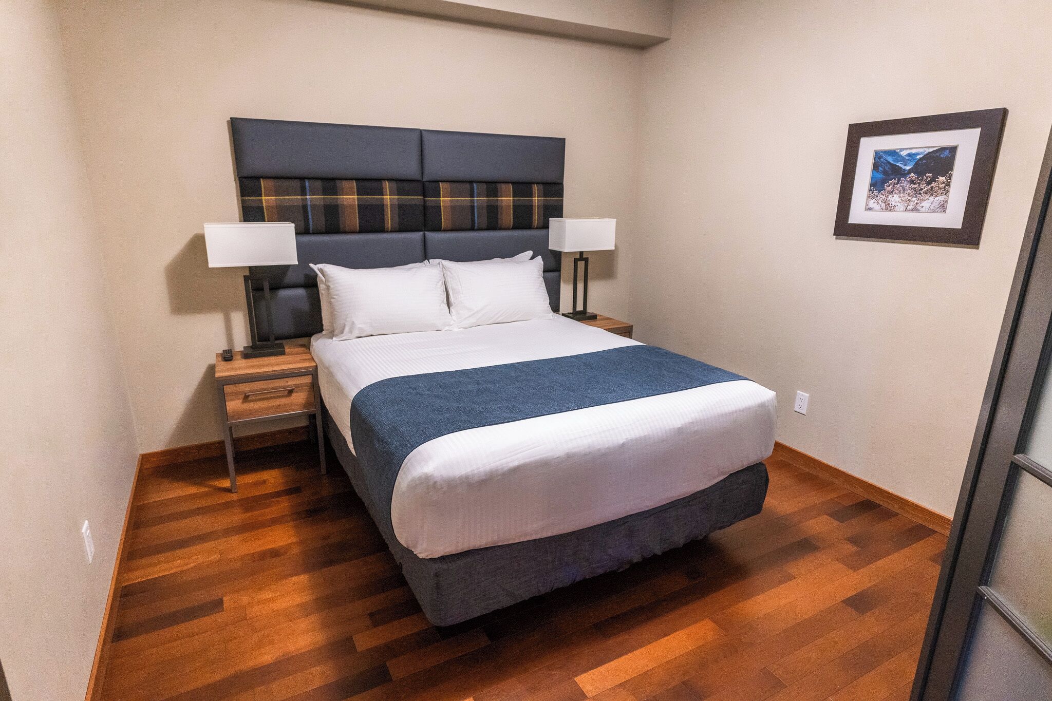 Una cama bien hecha con un cabecero oscuro, dos mesitas de noche con lámparas y un cuadro en la pared.