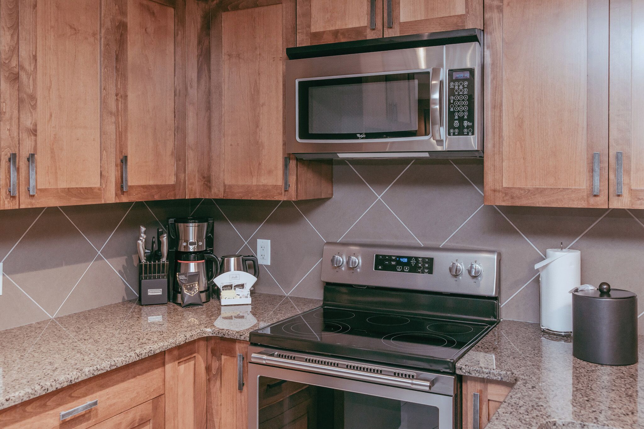 Una cocina con armarios de madera, microondas de acero inoxidable, cafetera, estufa y un cubo de basura.