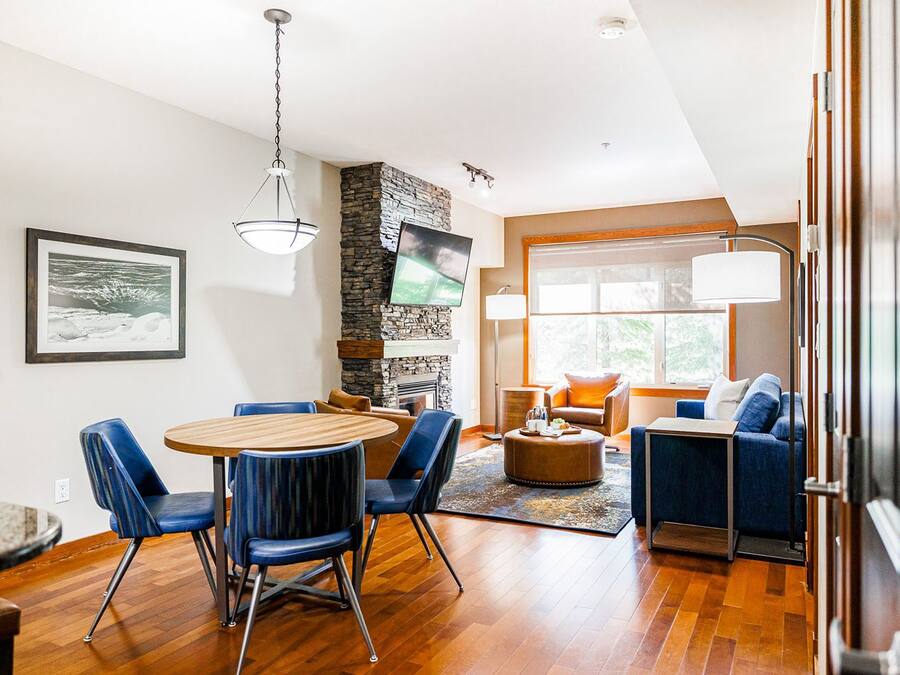 A modern living room with a wooden dining table, blue chairs, a stone fireplace, and a large window with a view.