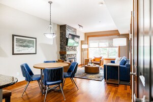 Una sala de estar moderna con una mesa de comedor de madera, sillas azules, una chimenea de piedra y un ventanal con vistas.