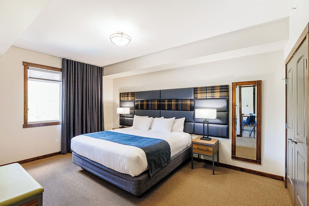 A hotel room with a large bed, a bedside table with a lamp, a mirror, and a window with curtains.