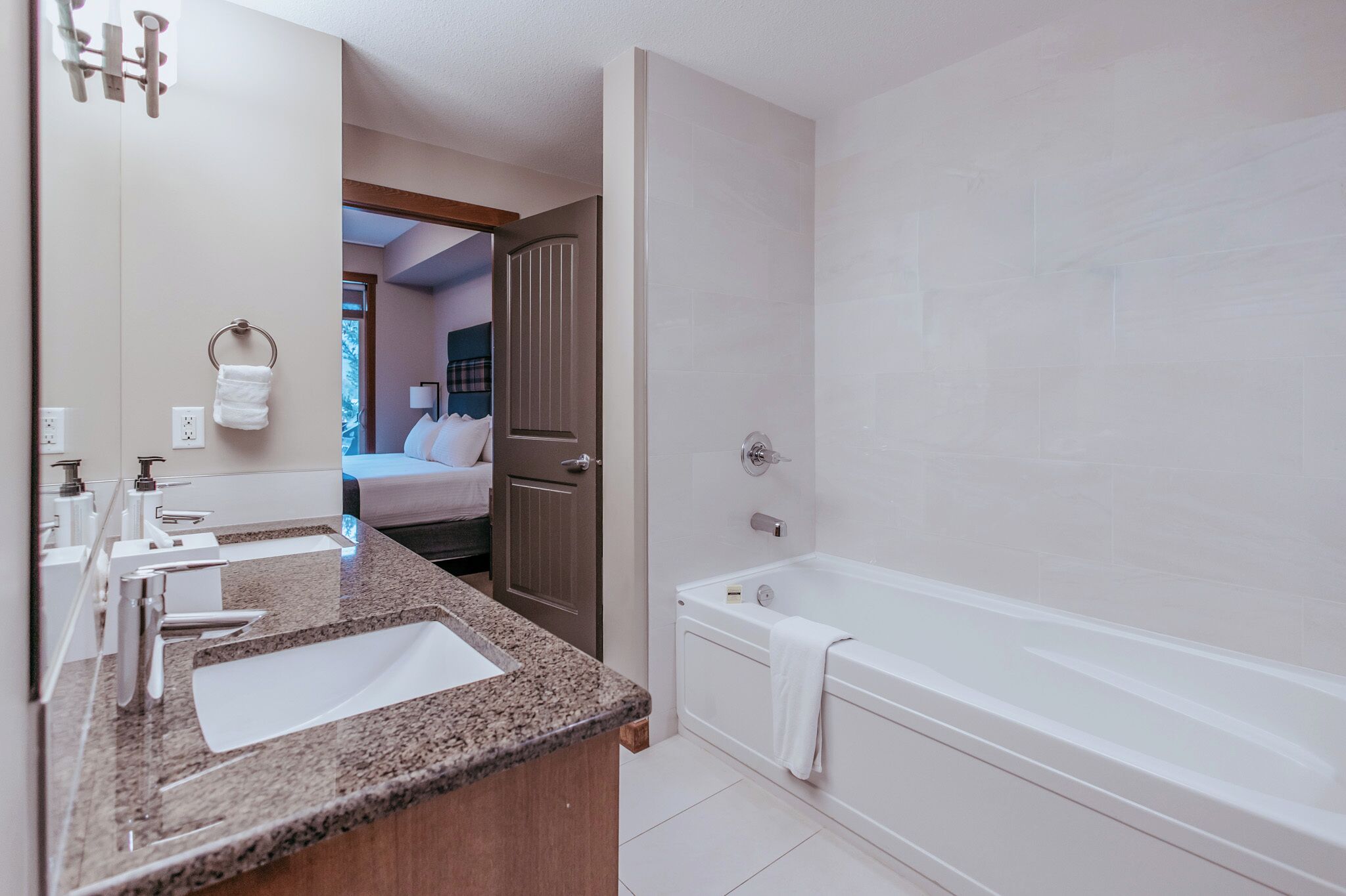 Un baño moderno con encimera de granito, una bañera y se ve un dormitorio con cama y ventana.