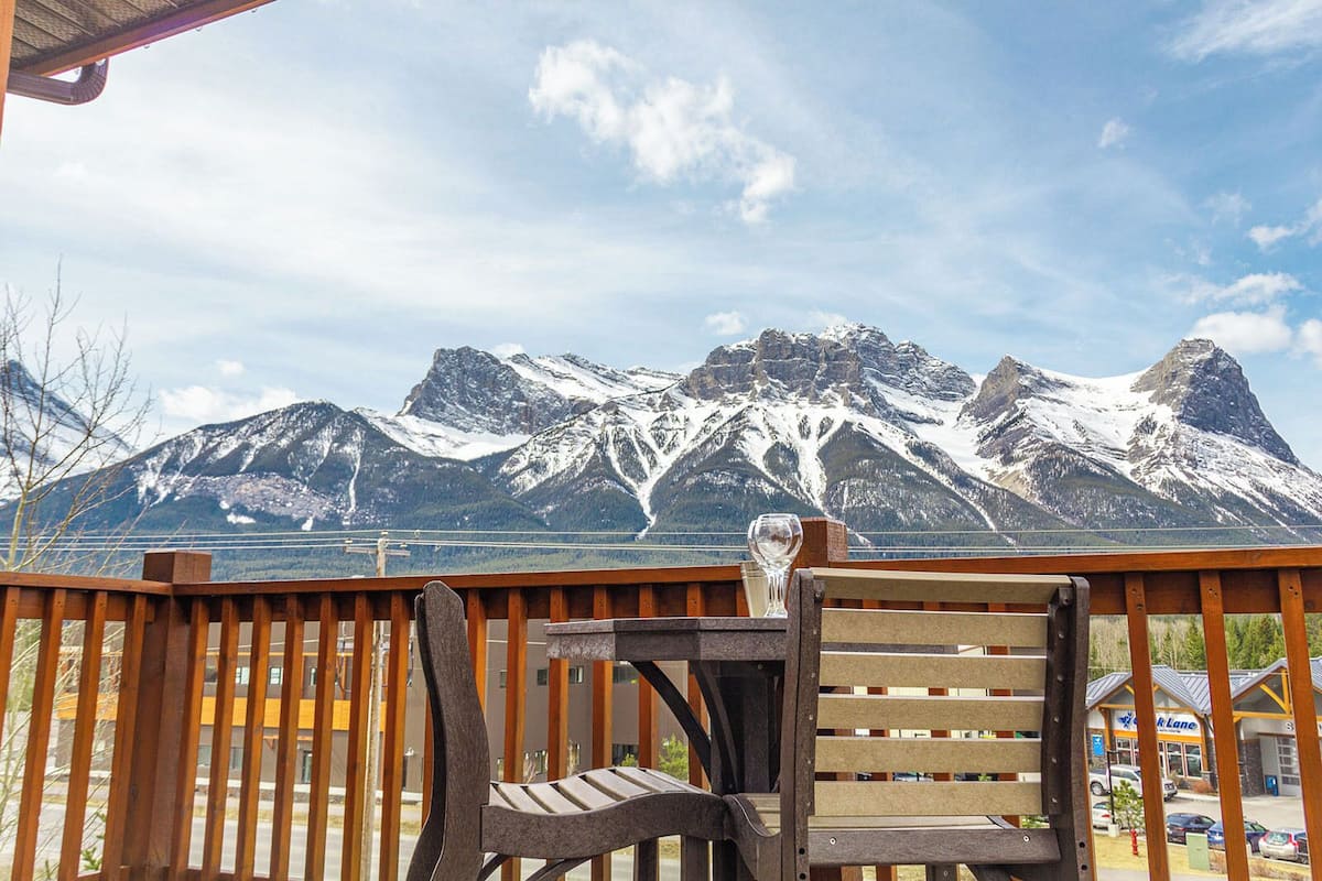 A wooden deck with a table and chairs, overlooking a mountain range with snow-capped peaks and a bridge in the background.