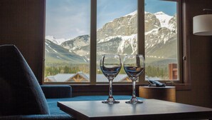 A room with a large window offering a mountain view, two empty wine glasses on a table, and a lamp on a side table. - Stoneridge Mountain Resort (Canmore)