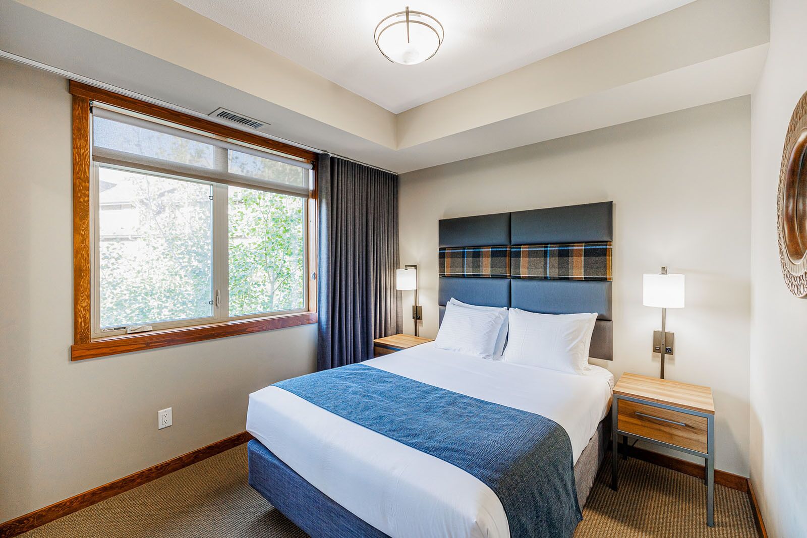 A hotel room with a large bed, a window with curtains, a headboard with a plaid pattern, bedside lamps, and a wall-mounted light fixture.