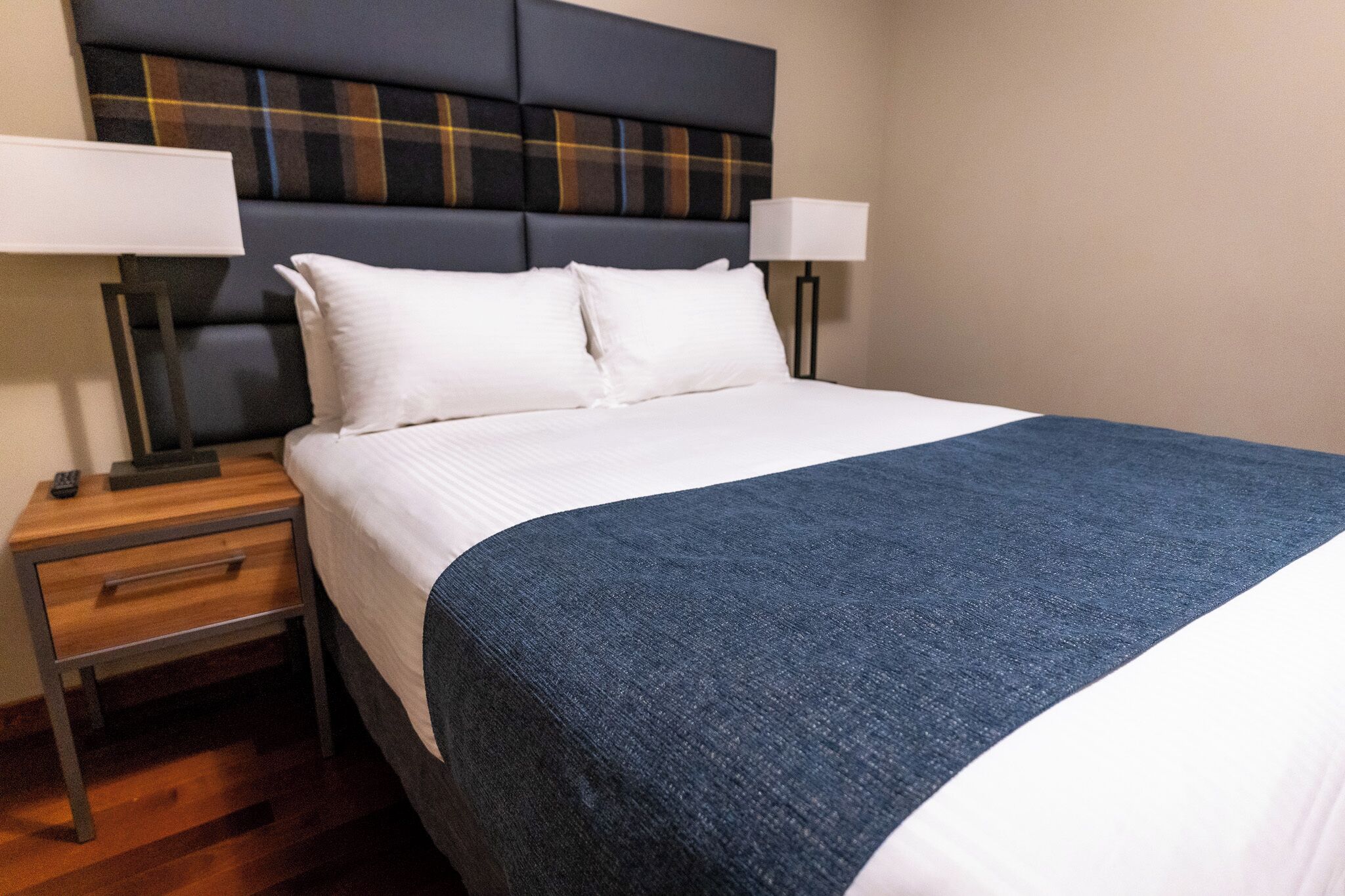 A neatly made bed with a blue blanket, two bedside lamps, a wooden nightstand with a drawer, and a headboard with a plaid pattern.