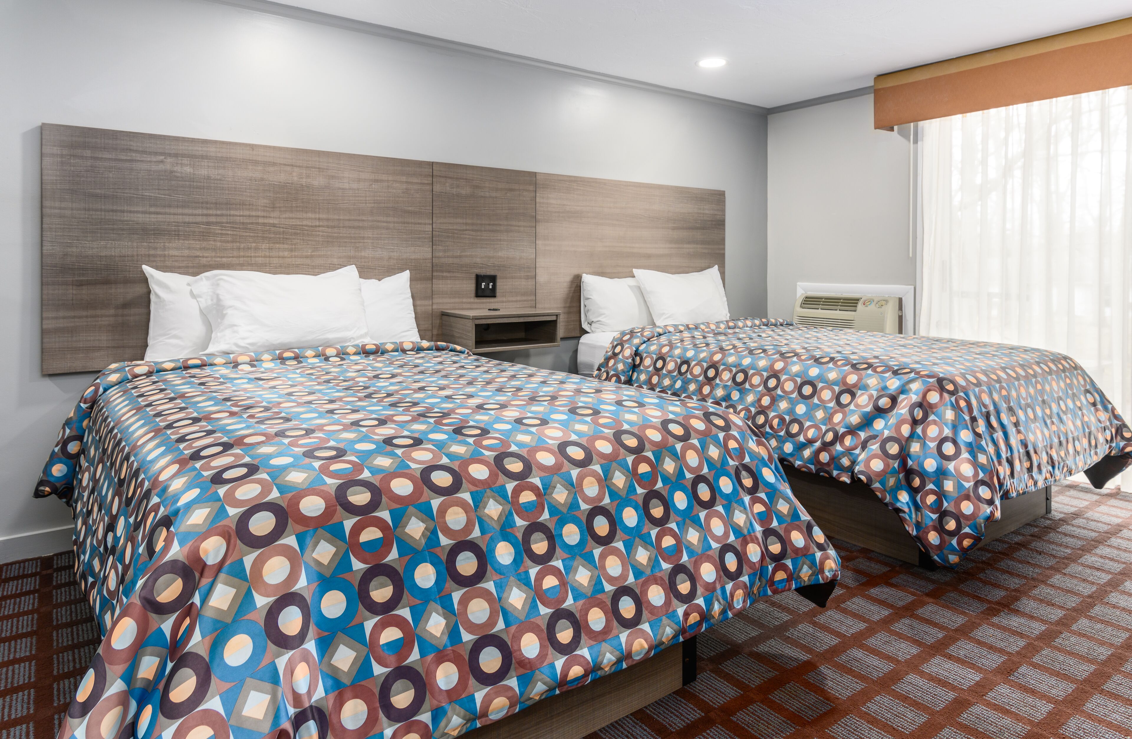 Habitación de hotel con dos camas, cabecero de madera, mesita de noche y ventana con cortinas.