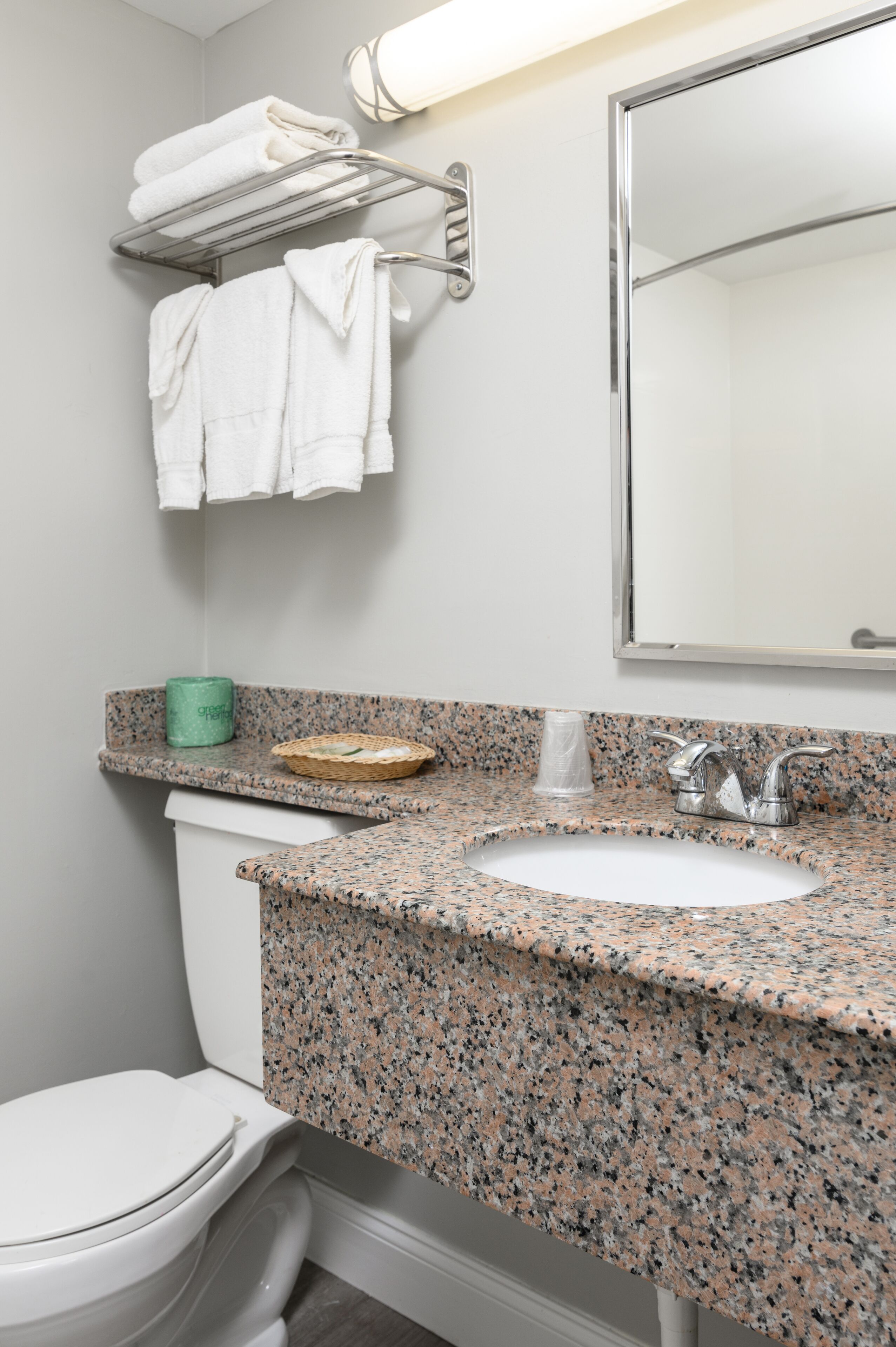 A bathroom with a granite countertop, a white sink, a towel rack with folded towels, a green towel dispenser, and a mirror.