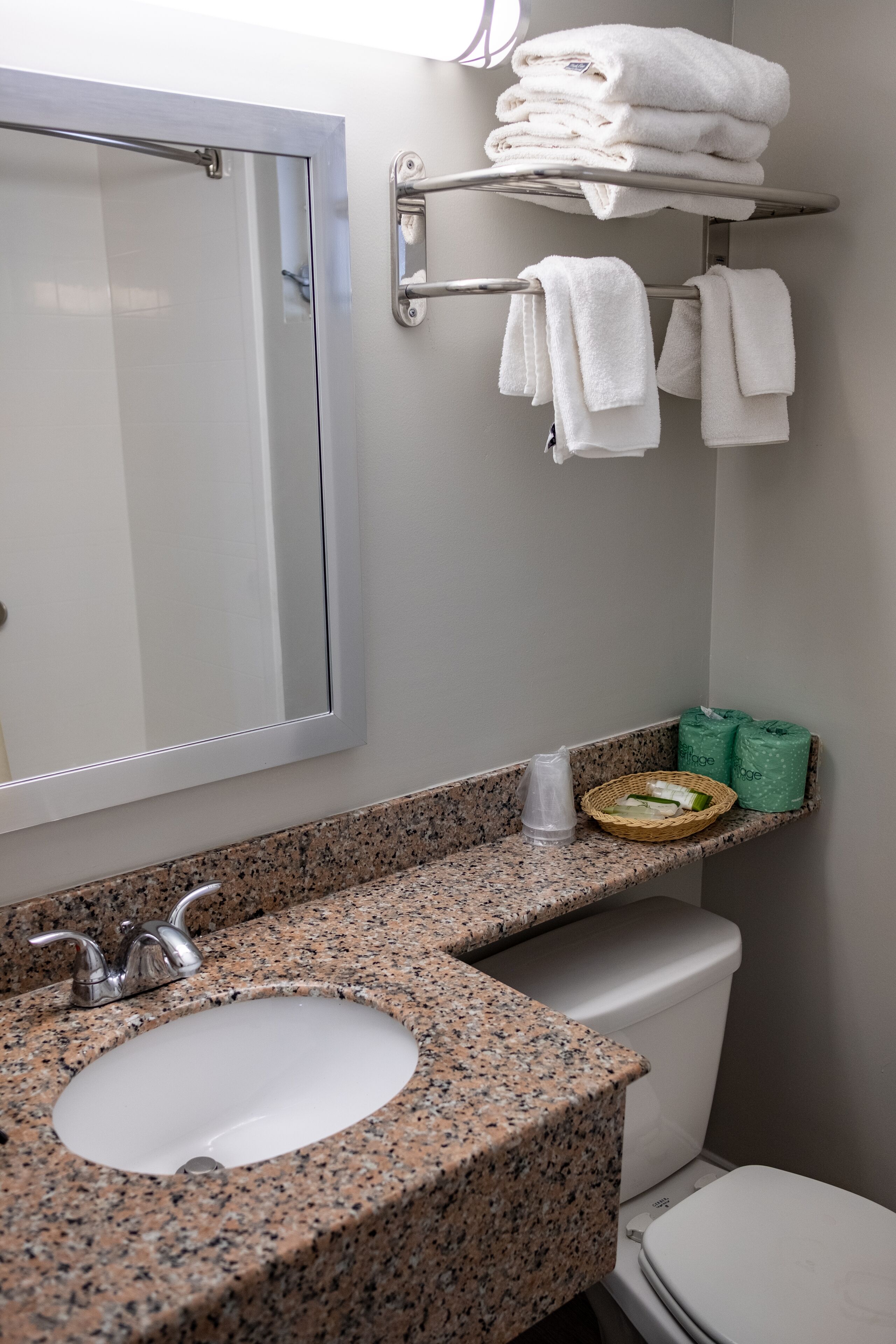 Casa de banho com bancada de granito, lavatório, sanita, porta-toalhas com toalhas dobradas, espelho e pequena prateleira com produtos de higiene pessoal.