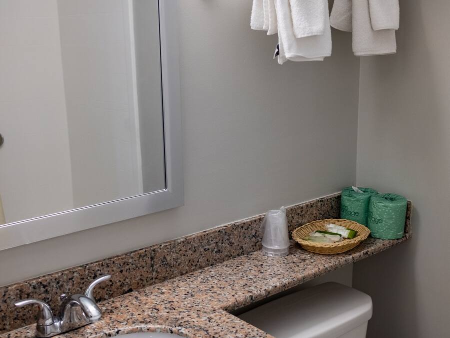 Casa de banho com bancada de granito, lavatório, sanita, porta-toalhas com toalhas dobradas, espelho e pequena prateleira com produtos de higiene pessoal.