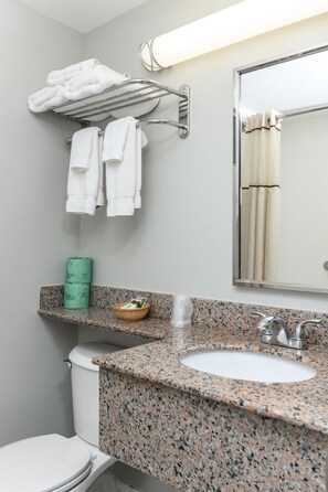 A bathroom with a granite countertop sink, a toilet, a towel rack with folded towels, a green towel dispenser, and a mirror.