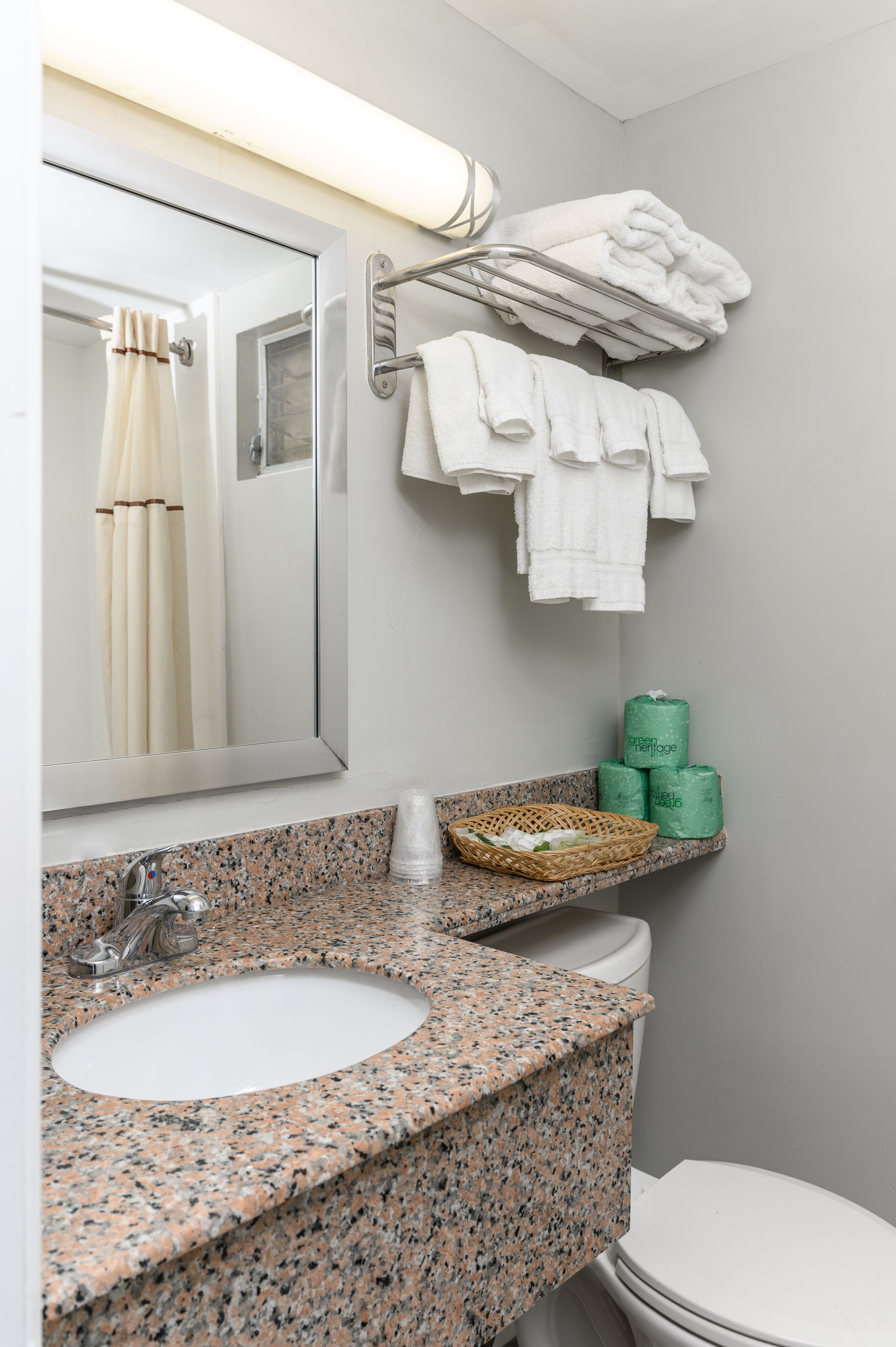 A bathroom with a granite countertop, a sink, a toilet, a towel rack with folded towels, a mirror, and a shower curtain.