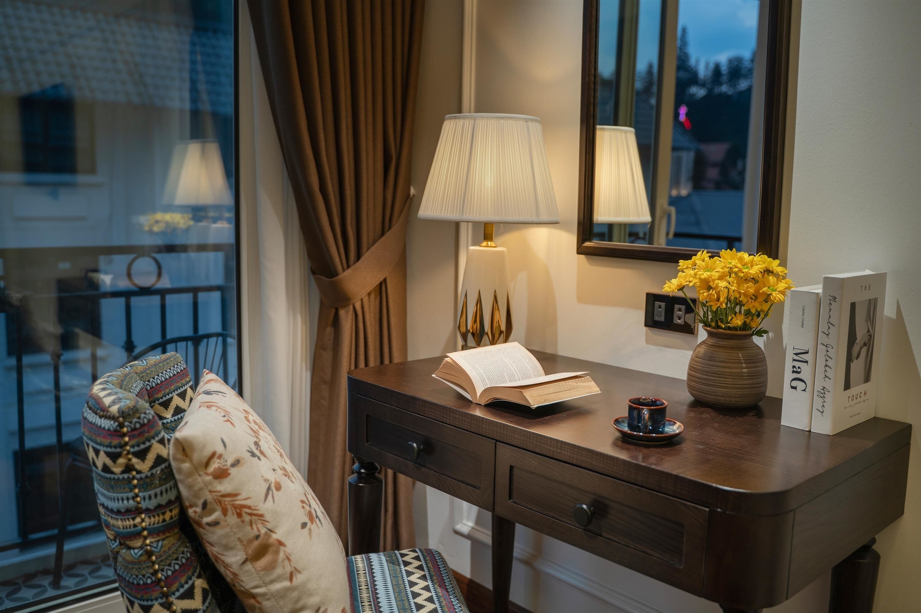 A wooden desk with a lamp, an open book, a vase with yellow flowers, and a framed picture.