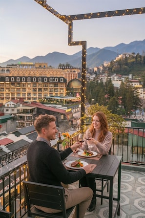 Um casal jantando em uma mesa ao ar livre com vista para a cidade, luzes de Natal e piso de azulejos.