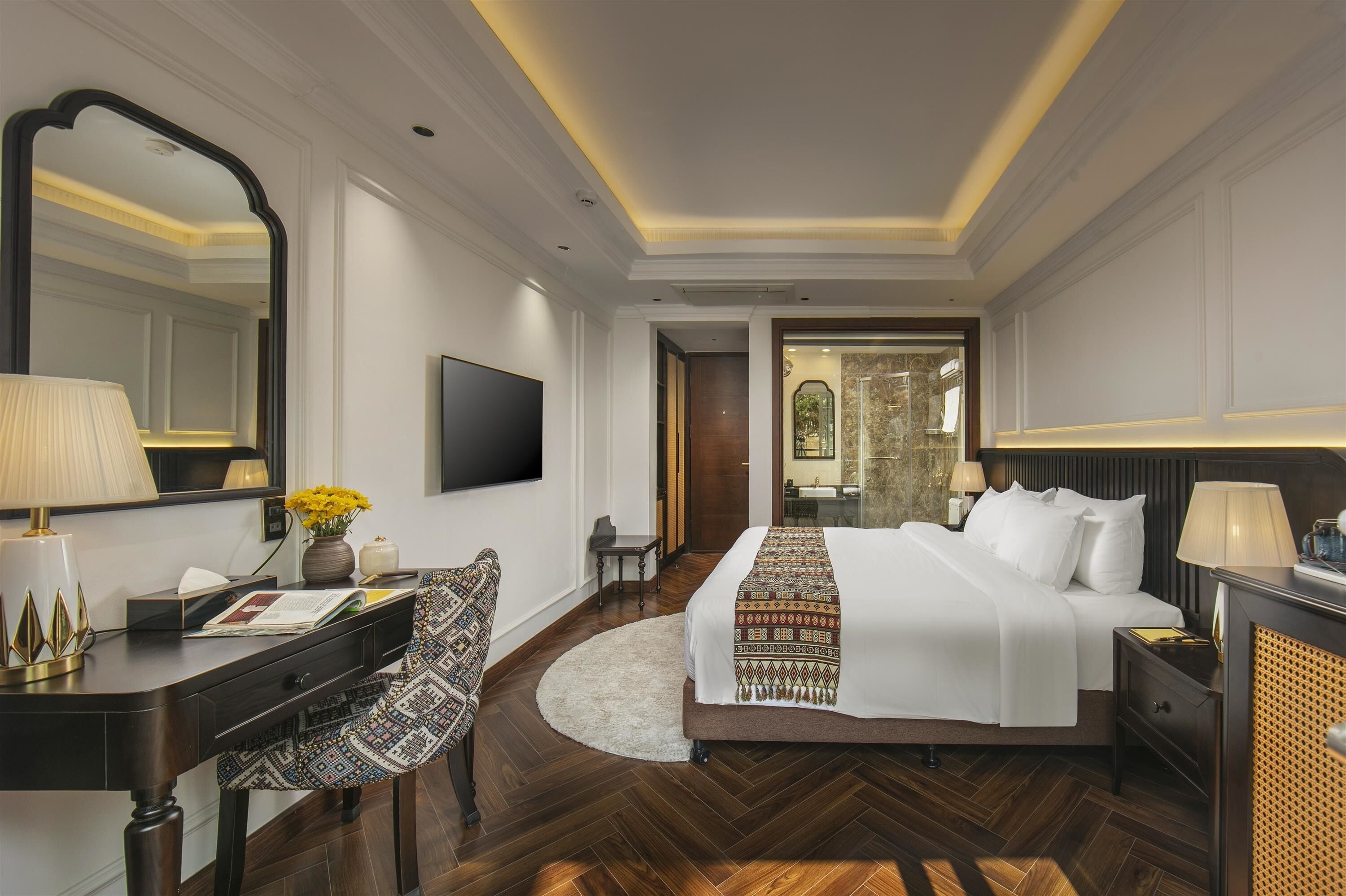 A hotel room with a large bed, a desk with a lamp, a chair, a TV, and a mirror.