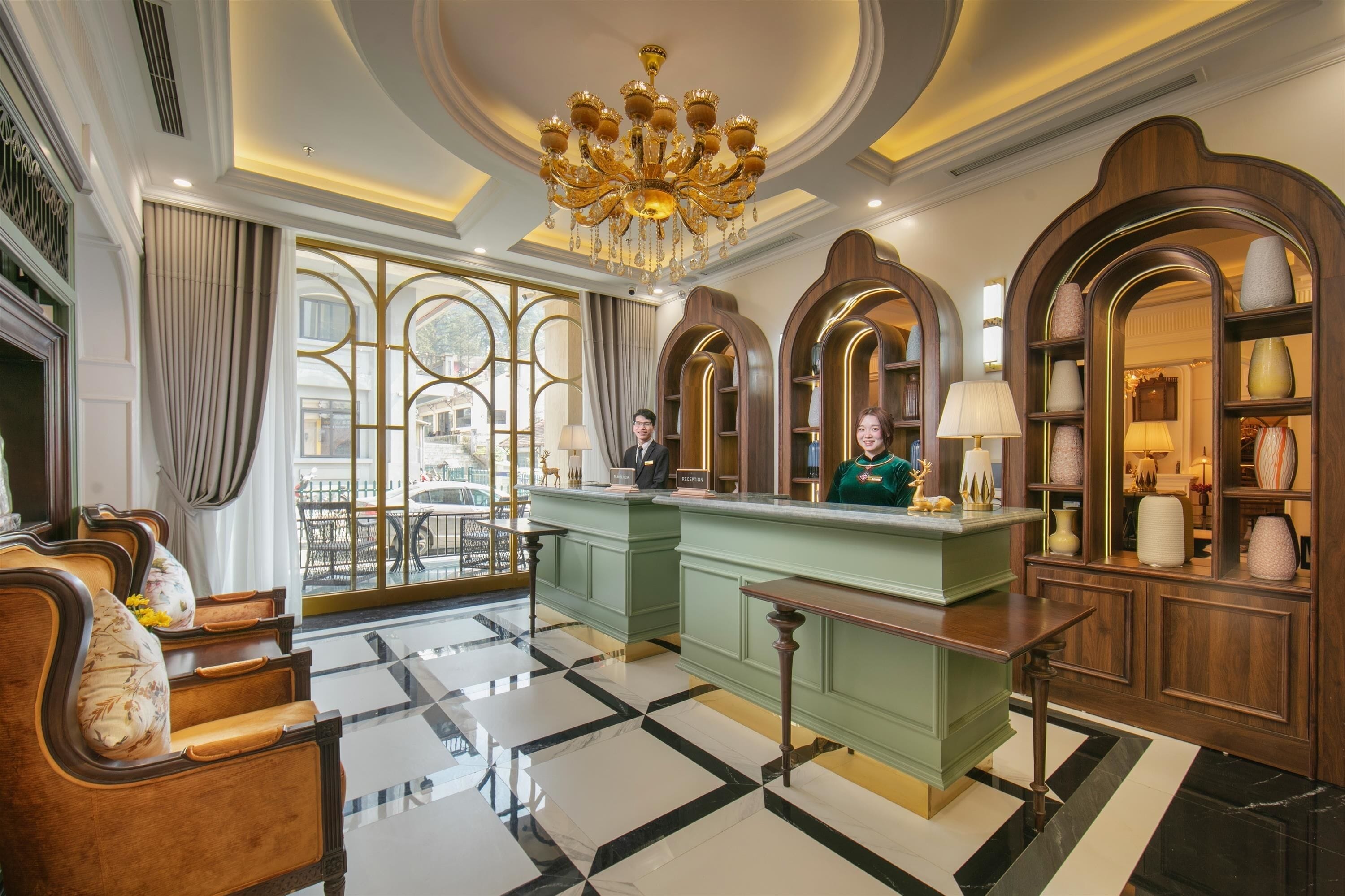 A hotel lobby with a reception desk, seating area, chandeliers, and decorative mirrors.