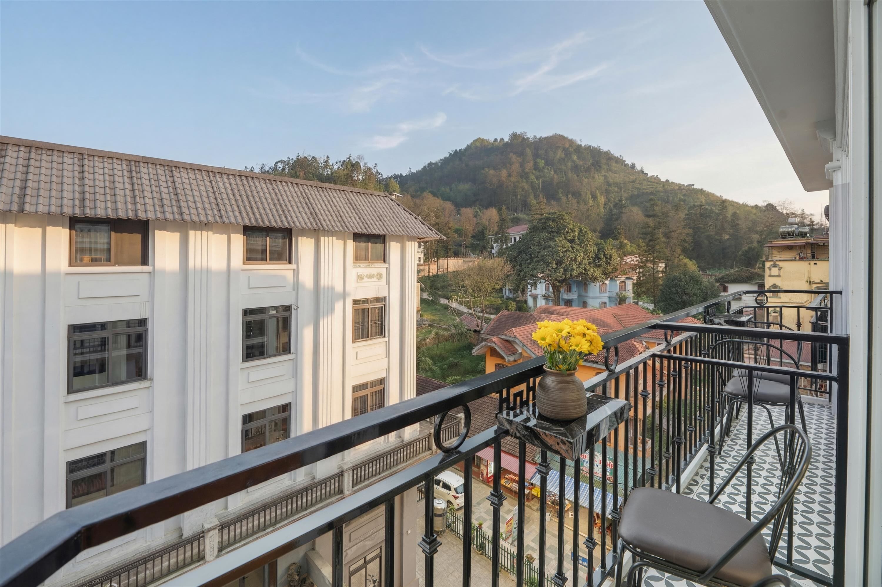 A balcony with a view of a building, a vase with flowers, and a chair.