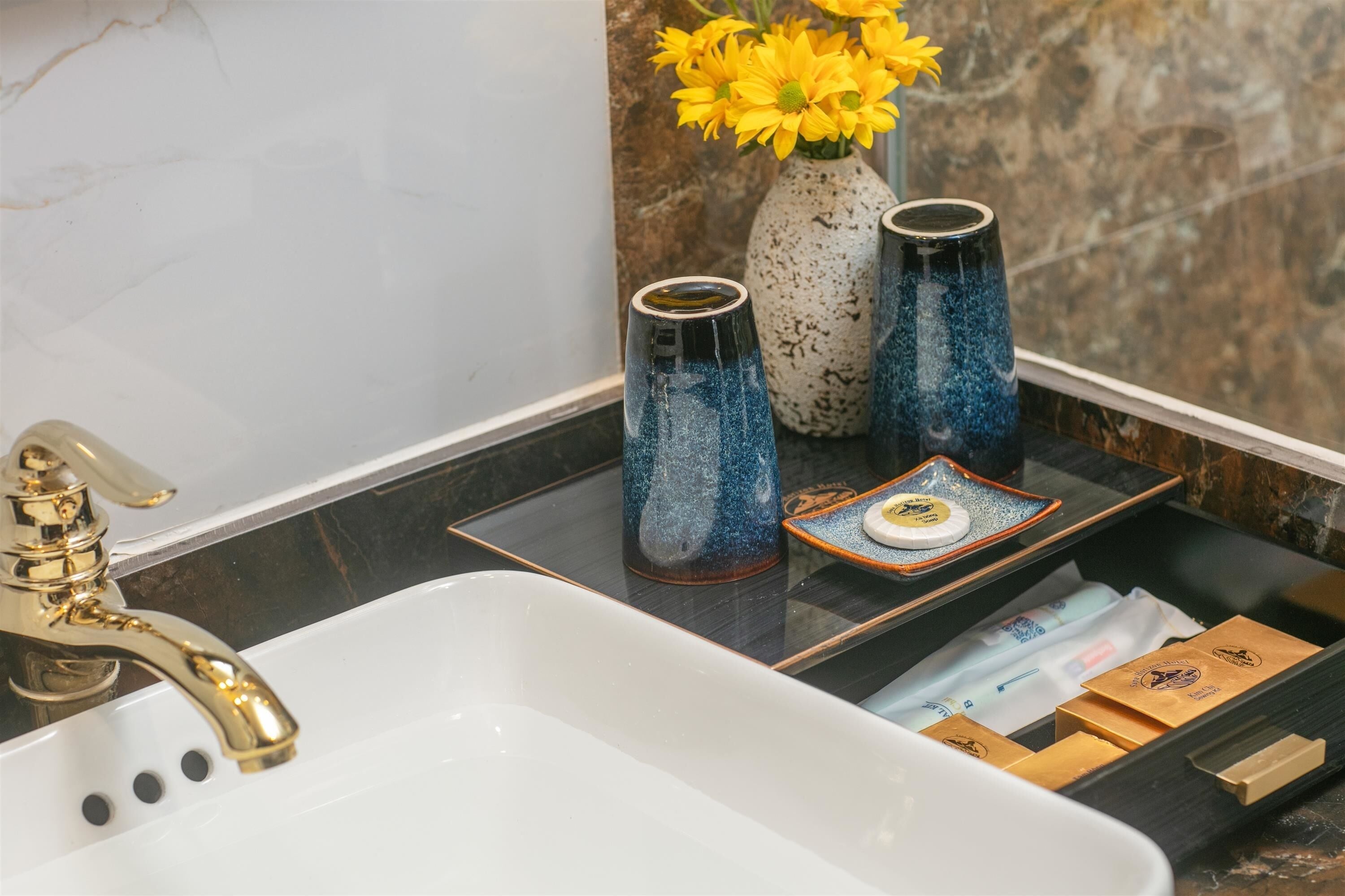Un baño con lavamanos blanco, grifo dorado y dos jarrones azules con motas, adornados con flores amarillas.