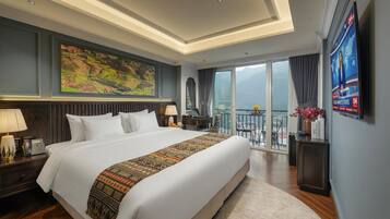 A hotel room with a large bed, a painting on the wall, a TV, and a balcony with a view.