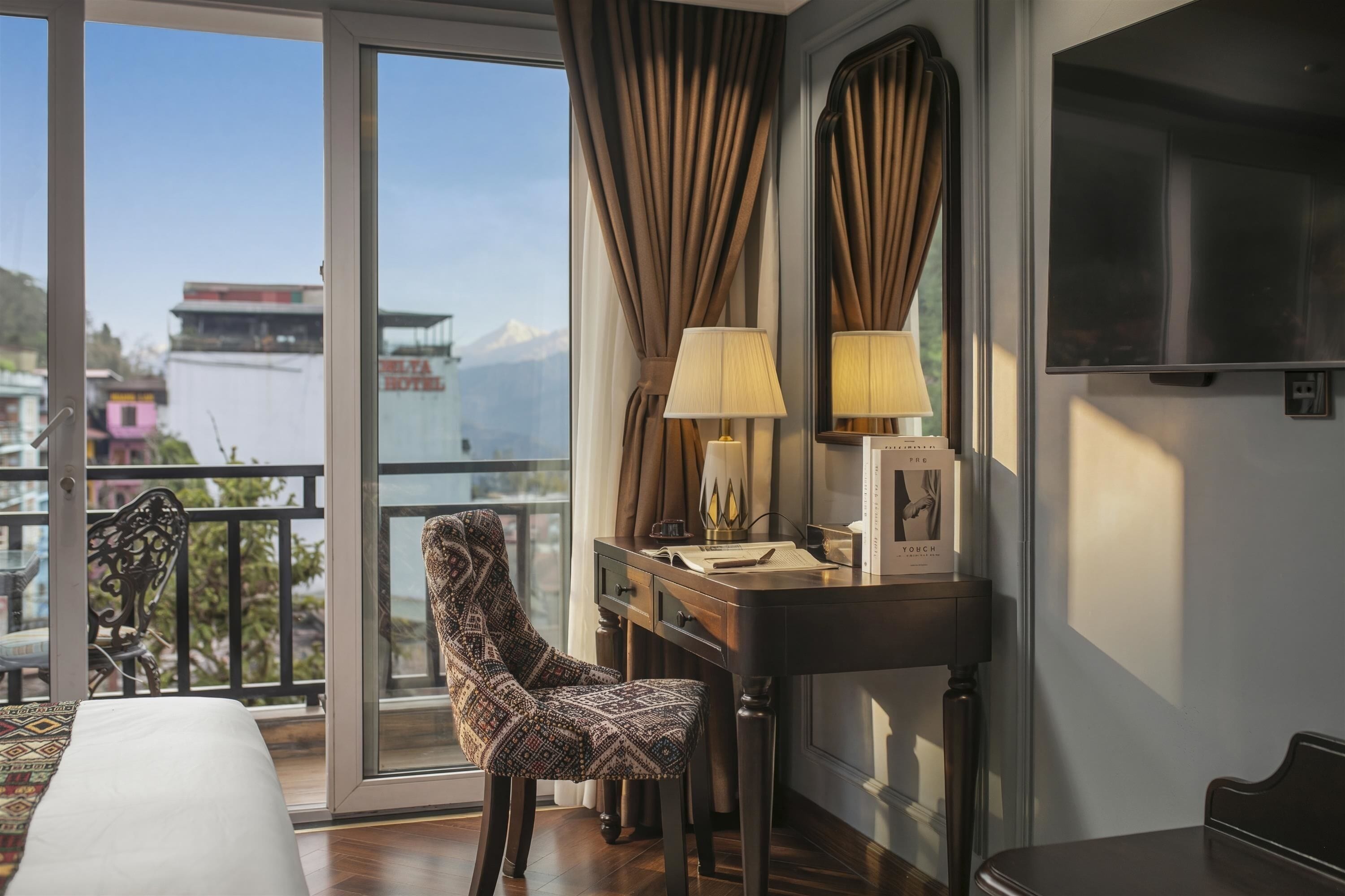 A hotel room with a bed, a desk with a lamp, a chair, a TV, and a balcony with a view of mountains and buildings.